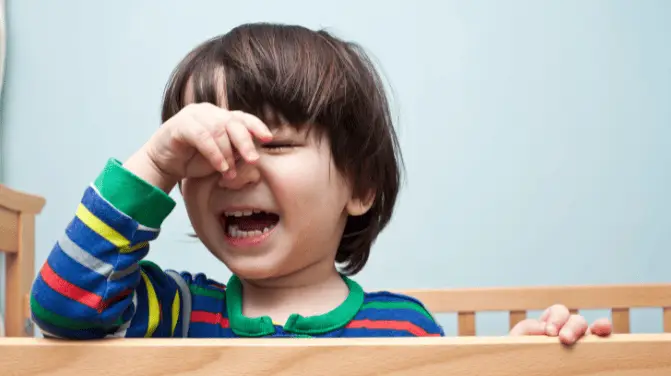 Baby in a playpen wearing a colourful top, crying while propped up