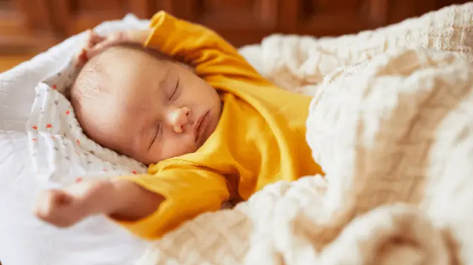 Little baby in a yellow sleepsuit sleeping under a soft white blanket