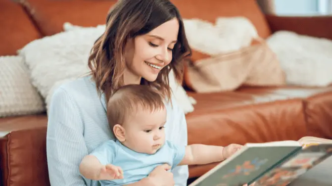 Young mother reading a book to her baby while leaning against a sofa