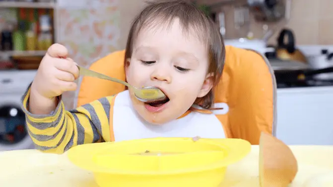 Baby eating soup alone from a yellow bowl